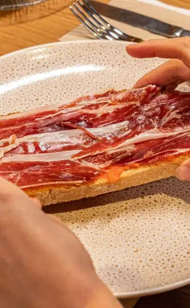 Slices of jamon iberico over tomato-rubbed toast on a speckled plate, a hand reaching for it