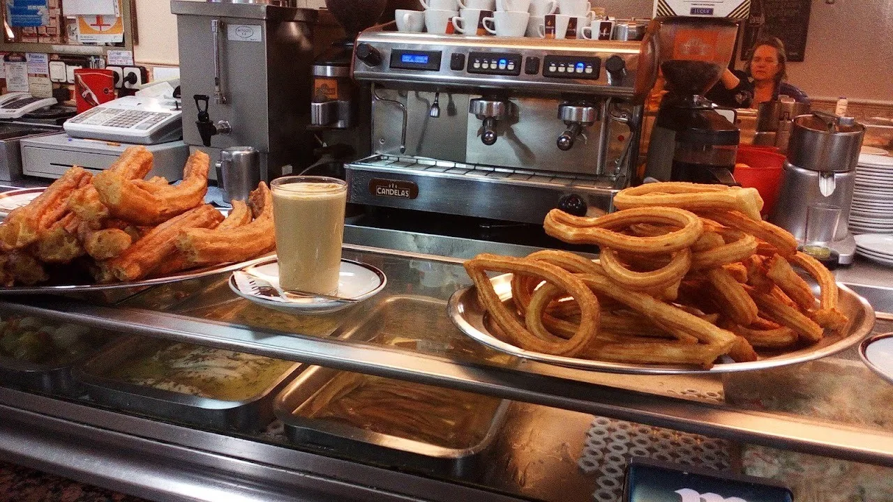 Trays of freshly fried churros and porras on a bar counter next to a glass of coffee
