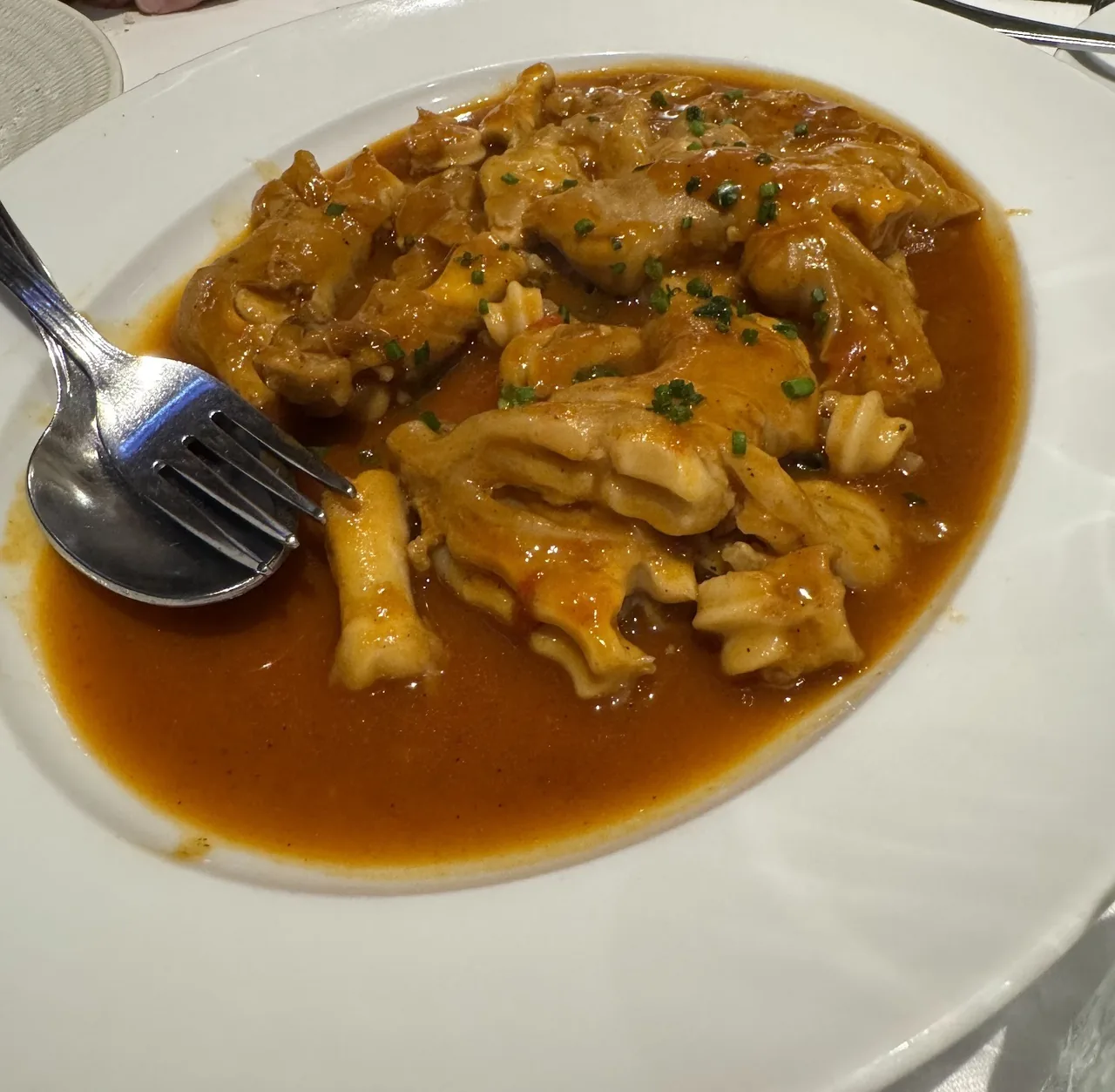 A white plate of tripe pieces in an orange-brown sauce with chopped chives and a fork