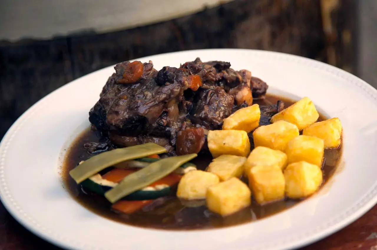 Braised meat in dark sauce with roasted potato cubes and grilled vegetables on a white plate