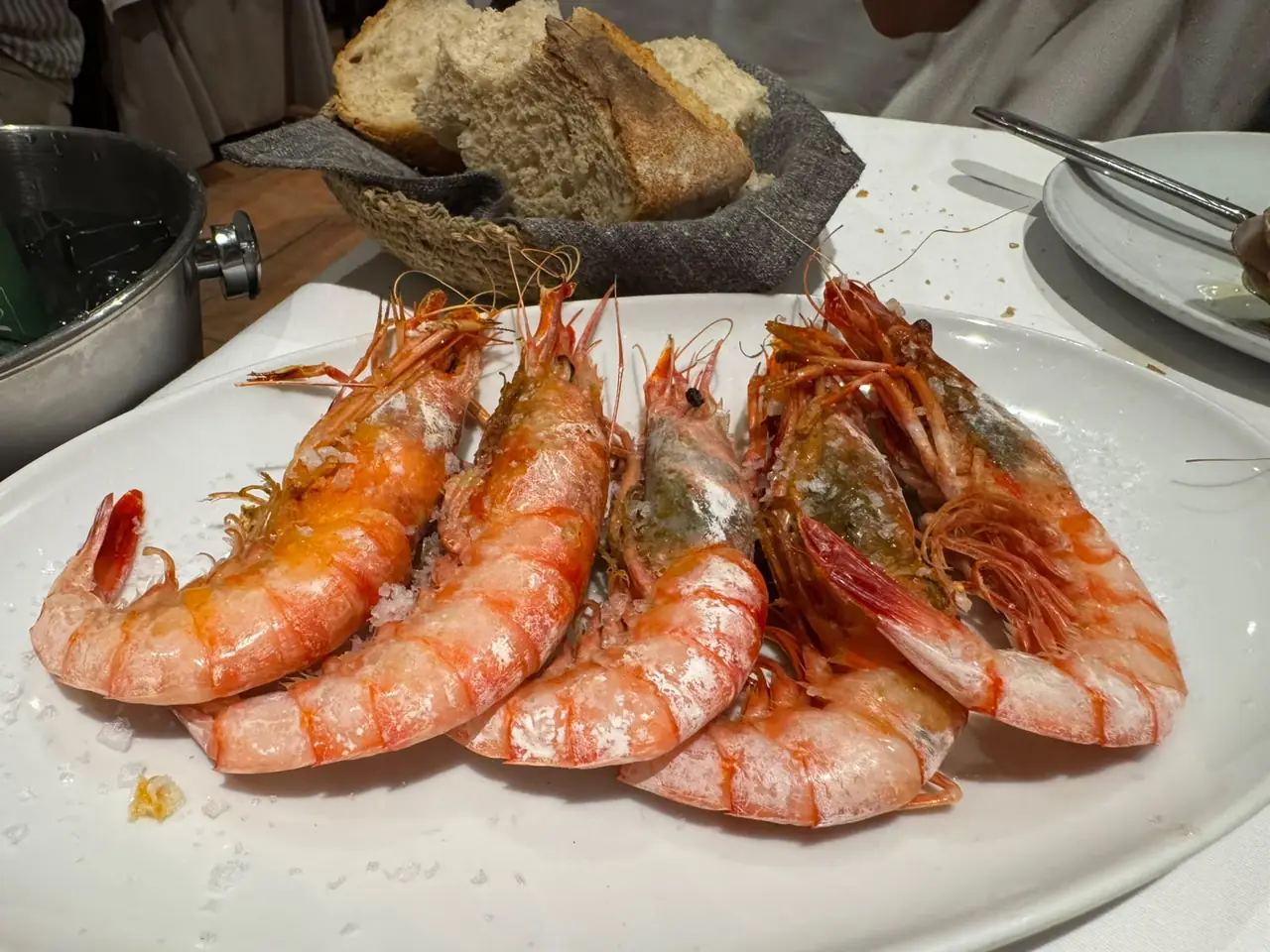 Whole grilled prawns with salt crystals on a white plate, bread basket behind