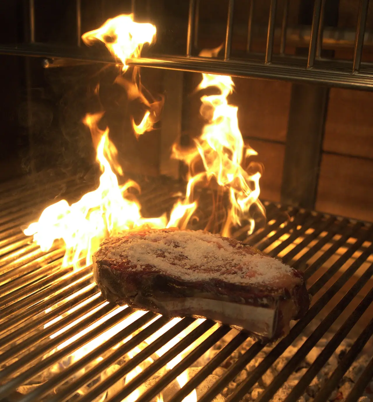 Thick salt-crusted steak searing directly over open flame on a wood-fired grill