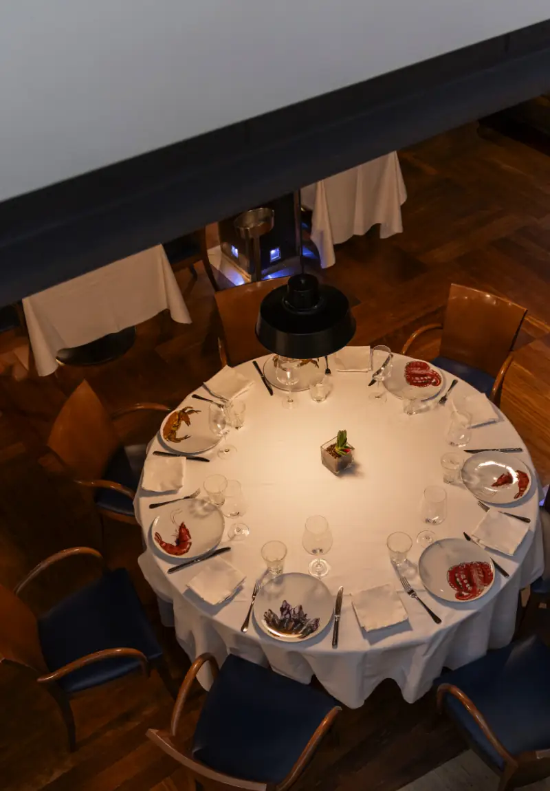 Overhead view of a round table set with white linens and plates of seafood starters including prawns and octopus, surrounded by dark blue chairs in a wood-floored dining room.