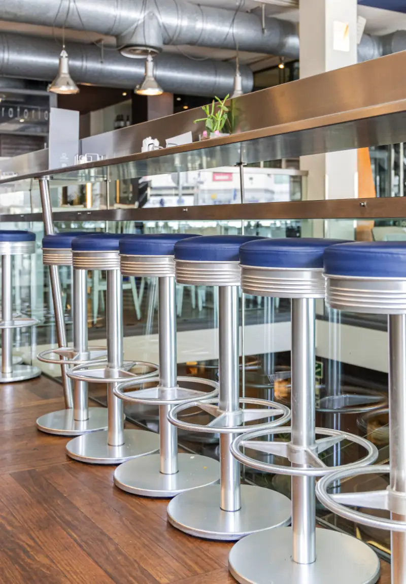 Row of blue-topped bar stools along a glass and steel counter, with exposed ductwork overhead.