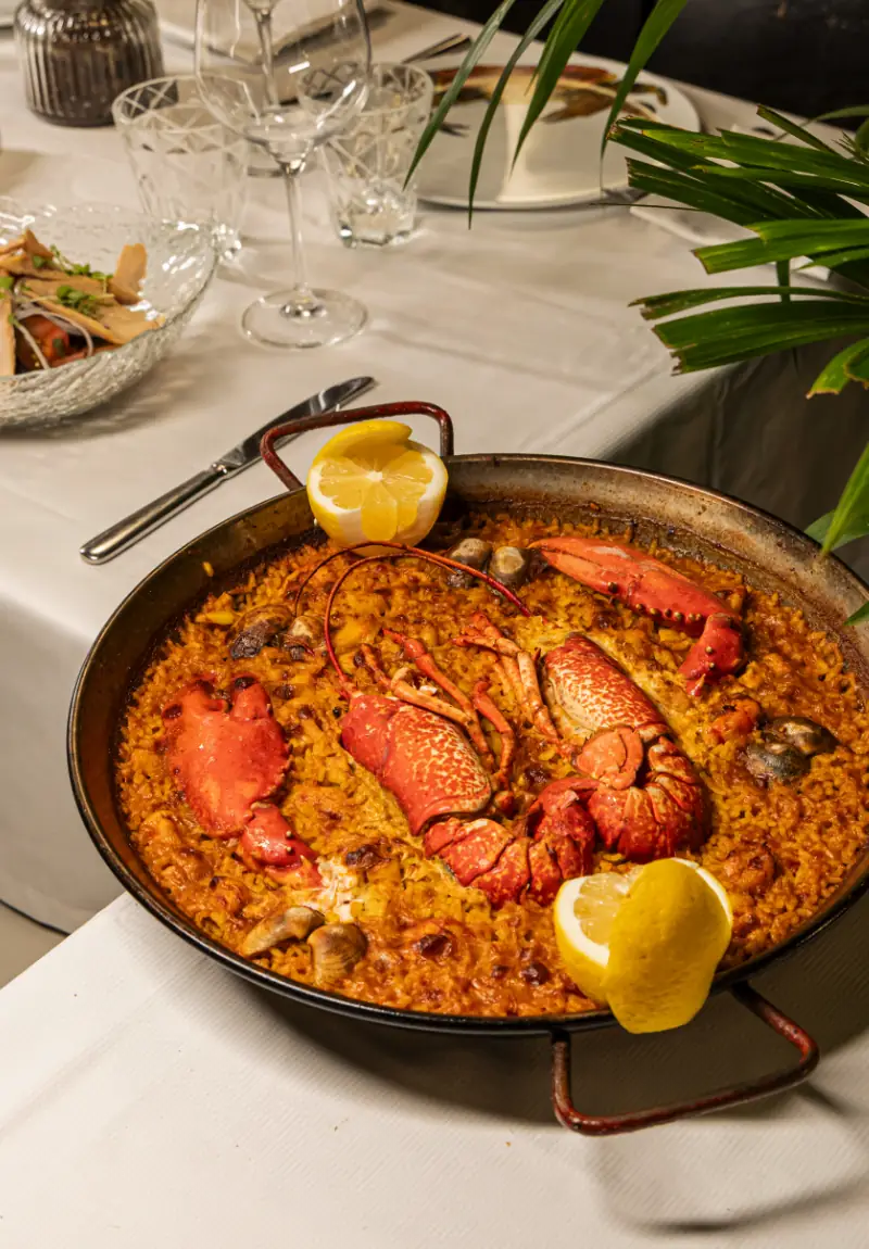 Seafood paella pan topped with whole lobster halves, clams and lemon wedges, set on a white-clothed table.