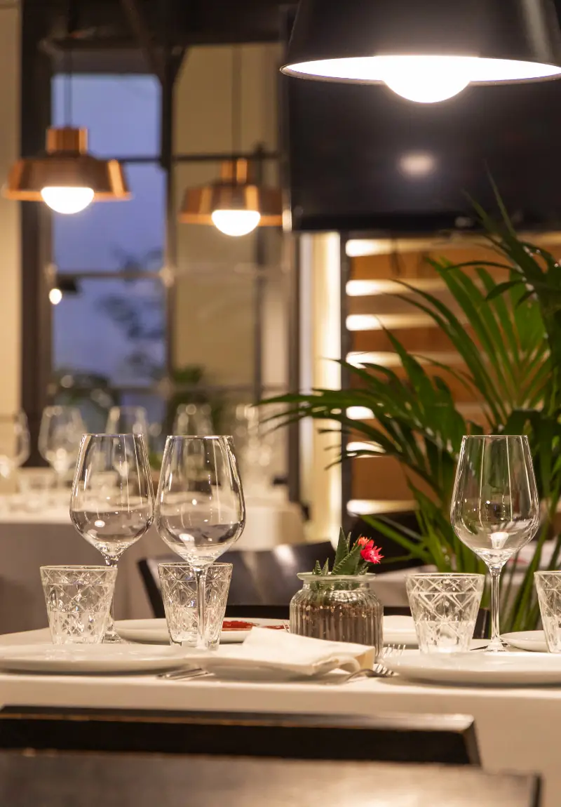 Table set with wine and water glasses under copper pendant lamps, with a potted plant and blurred dining room behind.