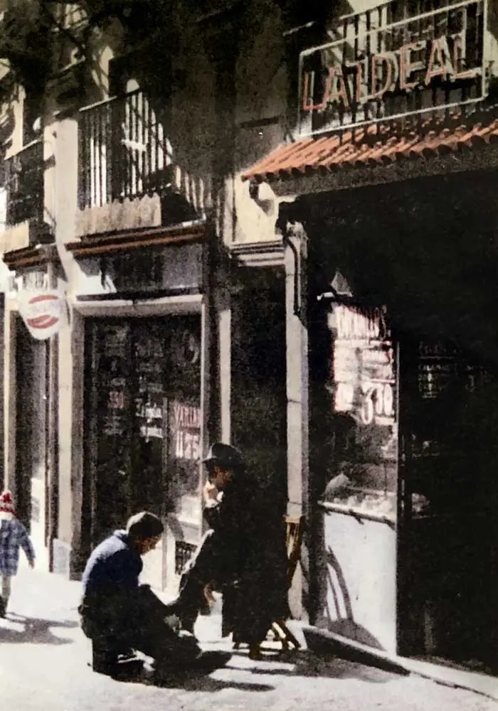 Vintage colorized street photo of the LA IDEAL storefront with its illuminated sign and a shoeshine scene on the pavement