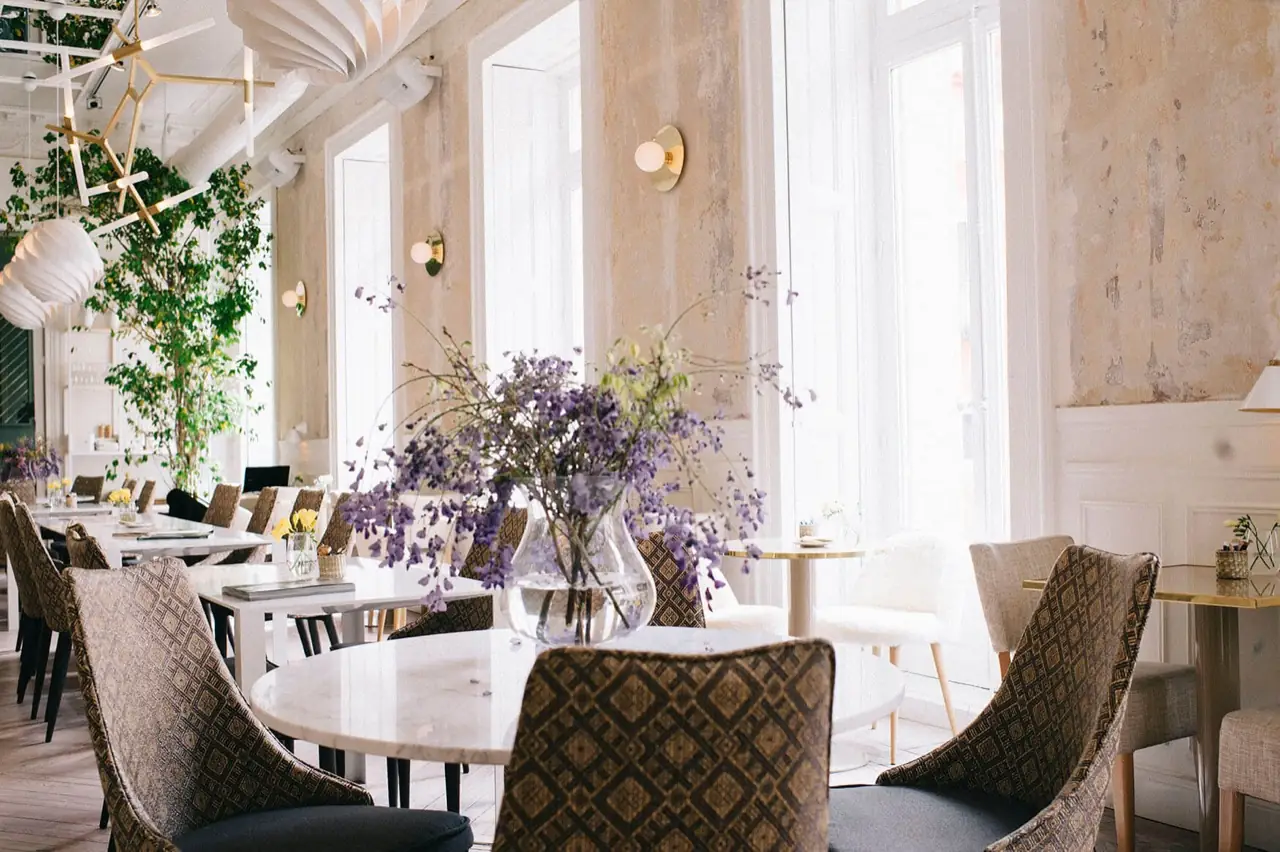 Bright dining room with high weathered plaster walls, tall arched windows, patterned upholstered chairs at marble tables, and a large glass vase of purple wisteria branches.