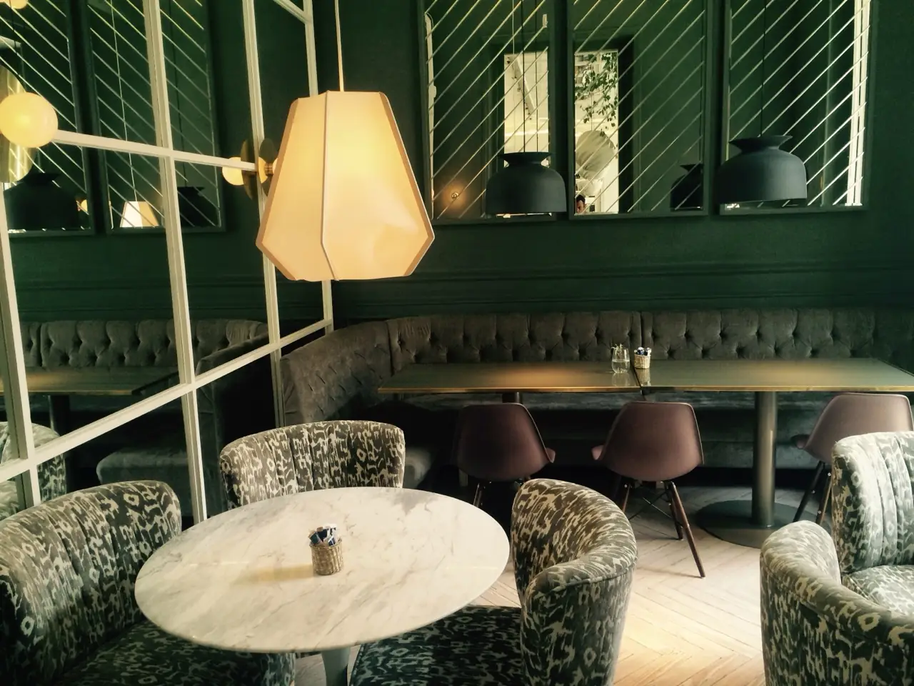 Dark green dining room with tufted velvet banquette seating, a geometric mirrored wall, a round marble table, and a geometric pendant lamp.