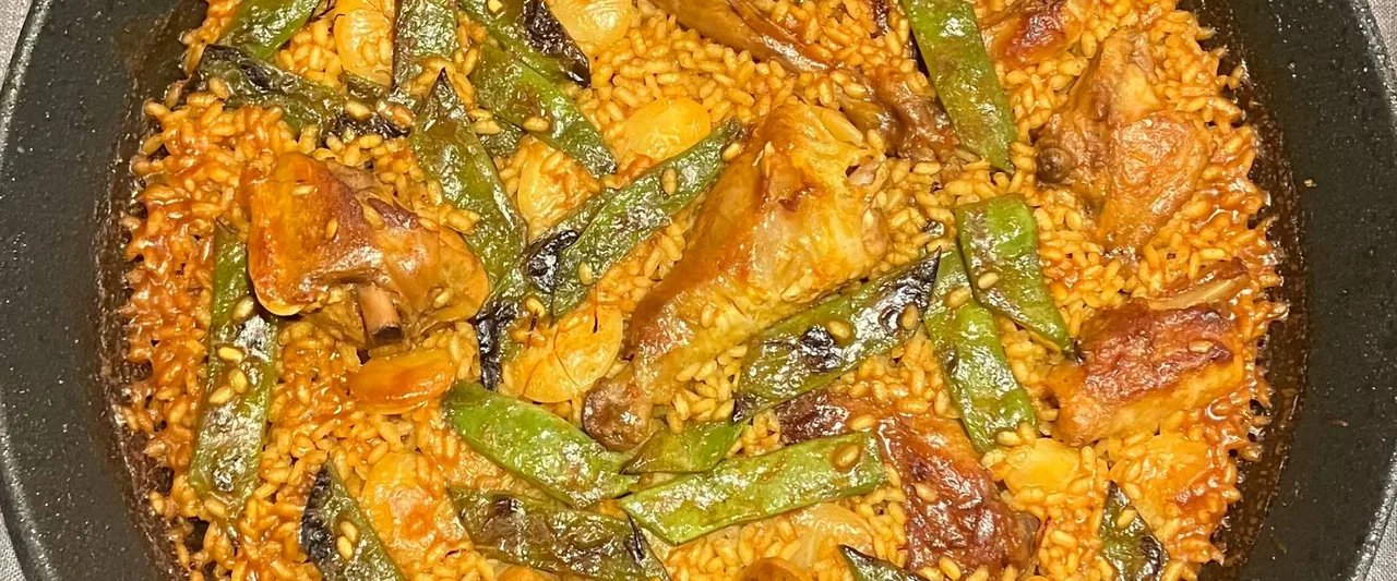 A wide crop of a paella in a dark pan: saffron-orange rice with flat green beans, browned chicken pieces and butter beans, crisped at the edges