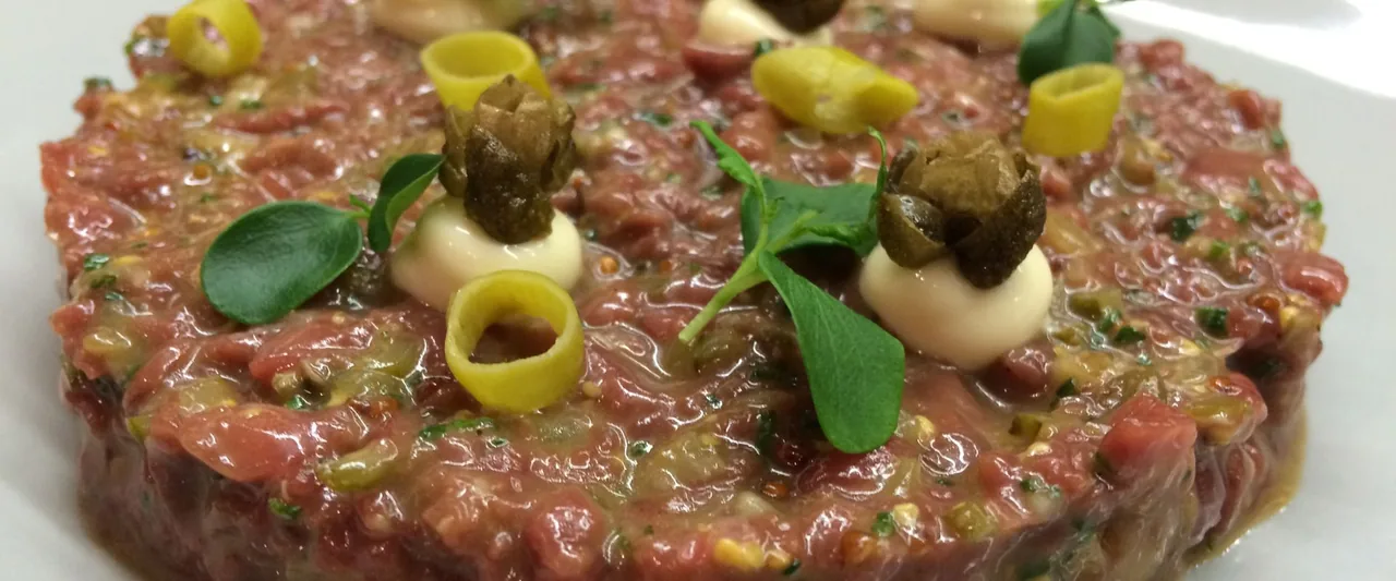A close crop of a beef tartare disc dressed with chopped gherkin, capers on dots of pale cream, curls of yellow pickled pepper and small green leaves