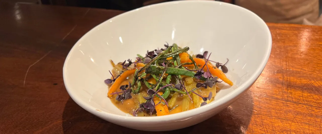 A white bowl of braised vegetables in a light broth, sliced carrot, celery and charred green beans under a scatter of purple micro leaves, on a dark wooden table
