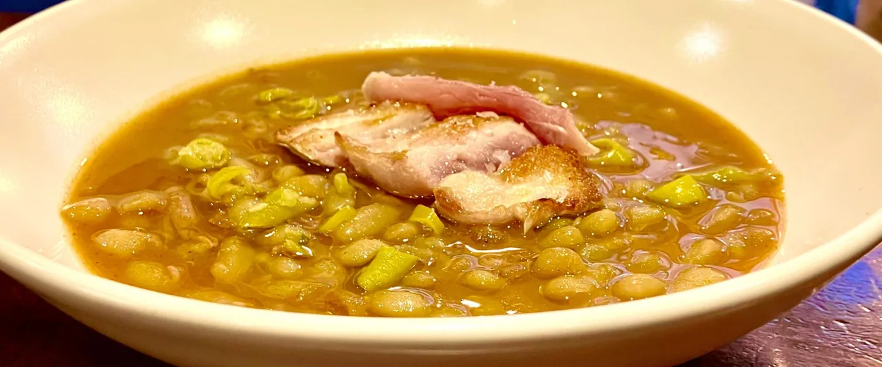 A white bowl of white bean and green bean stew in an amber broth, topped with slices of crisp-skinned pork belly