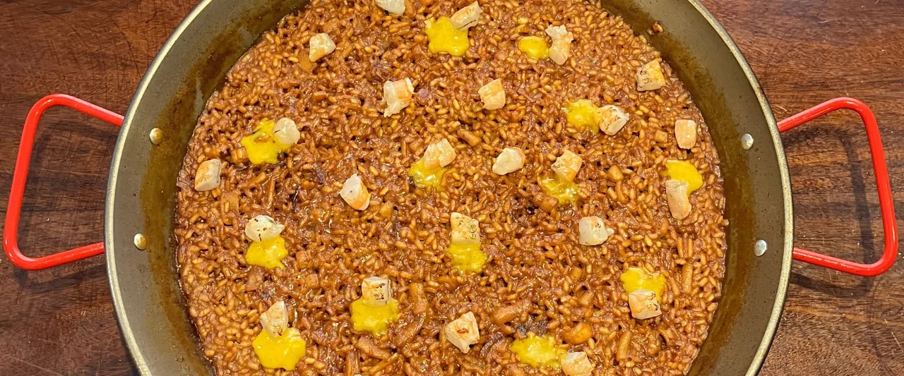 A red-handled paella pan of dark glossy rice dotted with pieces of white fish and small pools of yellow alioli, shot from directly overhead on a wooden table