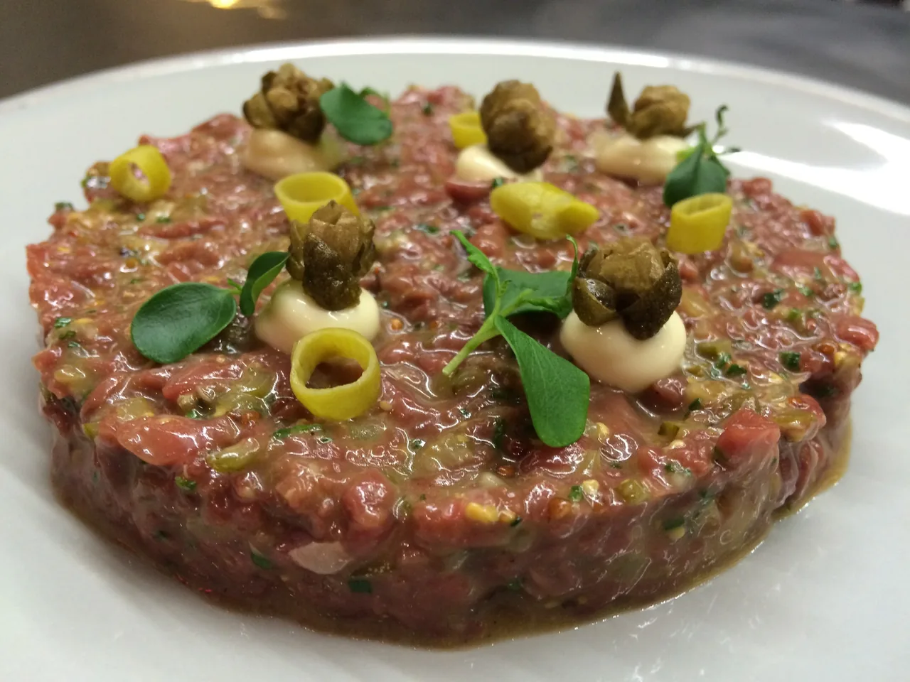 A round steak tartare topped with pickled vegetables and herbs on a white plate