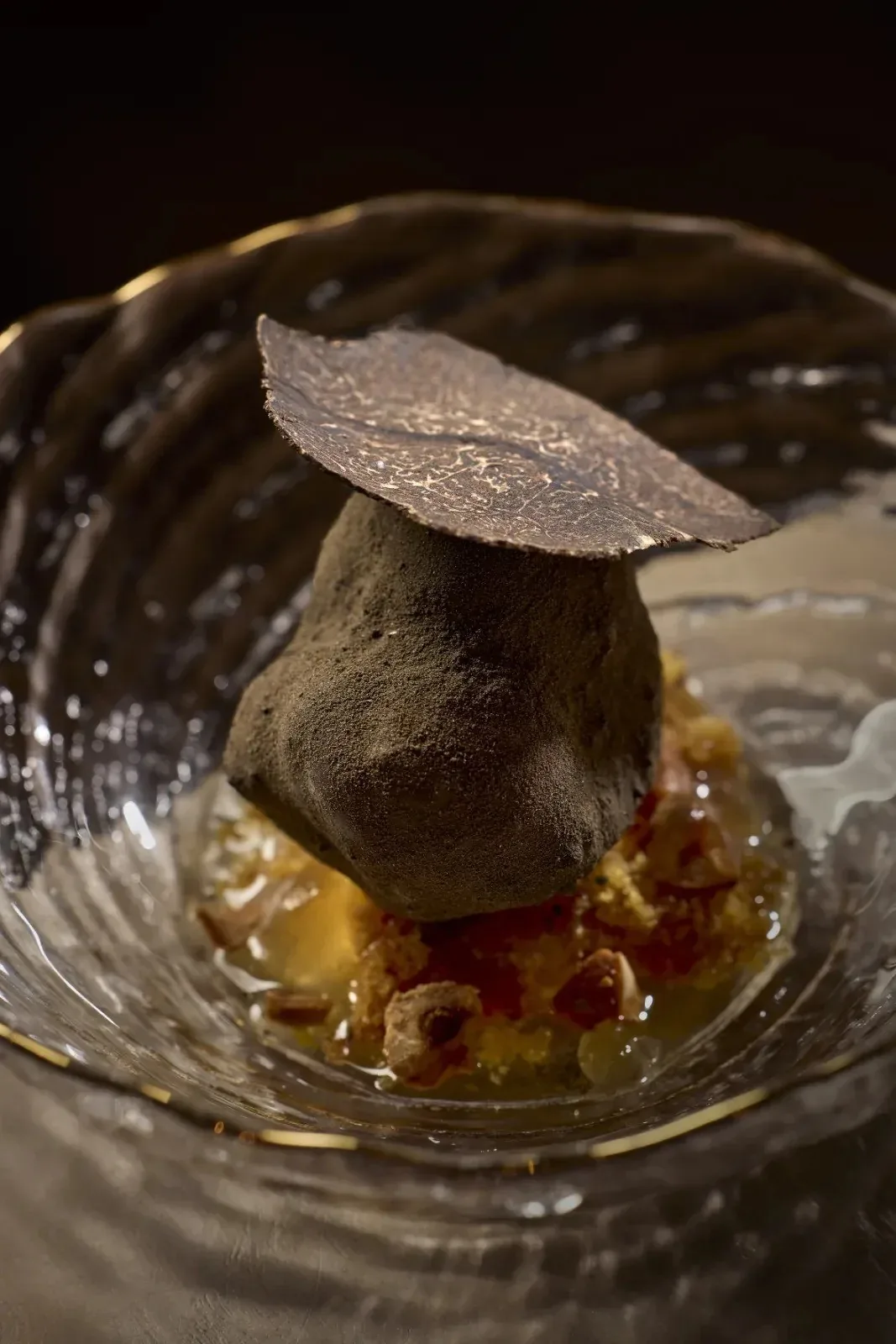 A dark-topped pebble-like dish under a thin crisp on a glass plate