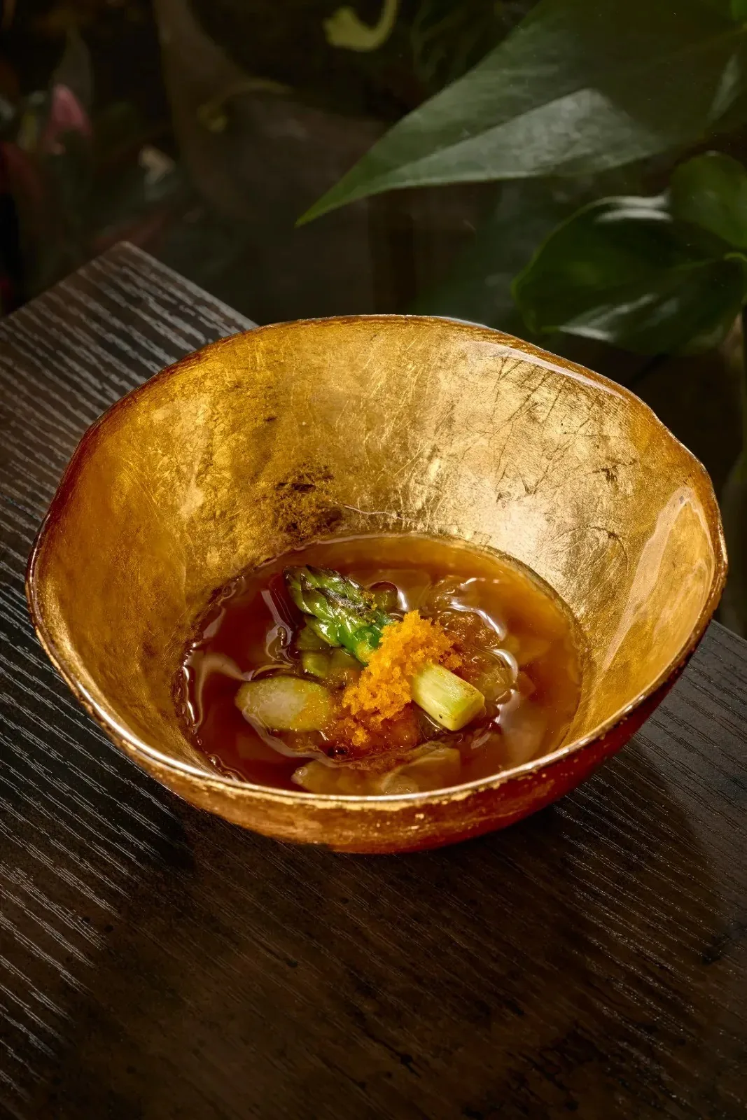 A gold-lined bowl holding a golden broth with small garnishes on a wooden counter