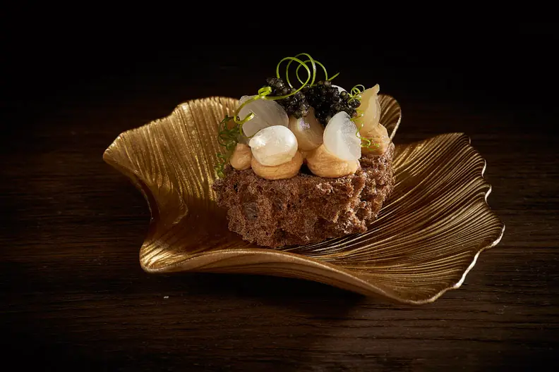 Small bite on a dark crumb base topped with caviar, pearl onions and herb curls, on a gold leaf-shaped dish