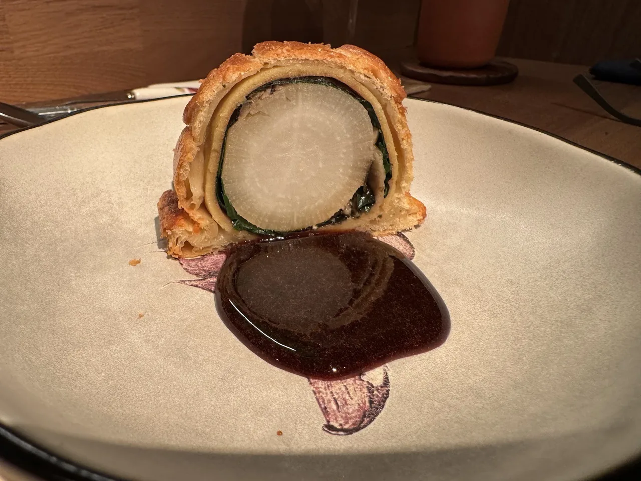 A pastry-wrapped slice with a round white vegetable centre beside a dark glossy sauce on a cream plate