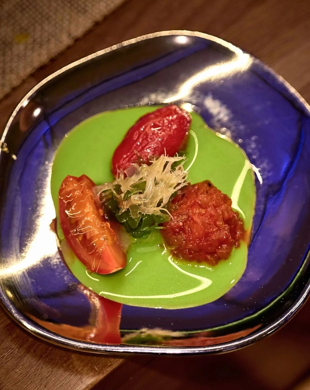 A blue glazed plate with a green sauce and red vegetable pieces topped with fine shredded garnish