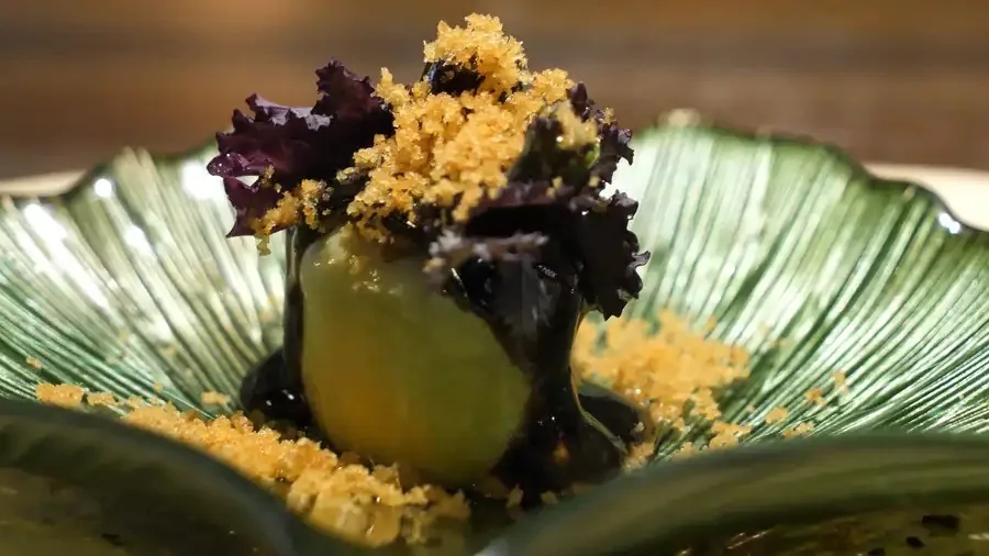 A dark leafy garnish with golden crumb on a round green vegetable, served on a green ribbed plate
