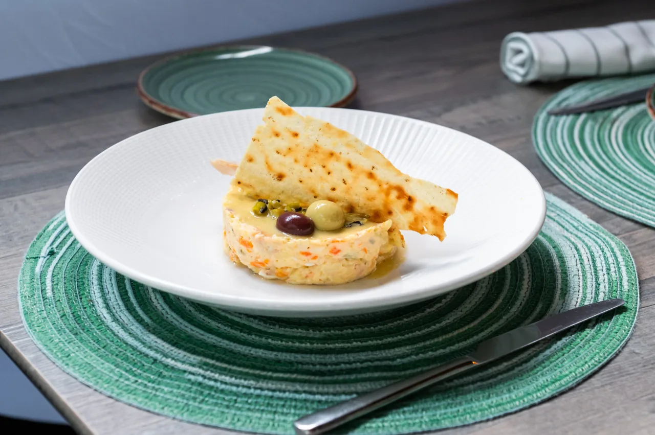A ring-moulded ensaladilla rusa topped with a black and a green olive and a shard of thin toasted flatbread, on a white plate over a green woven placemat