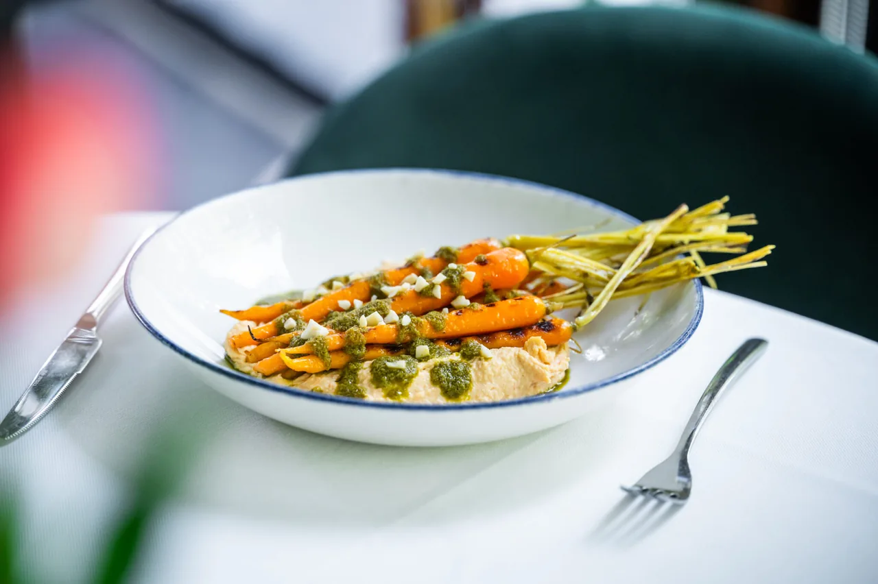 Whole roasted baby carrots with their tops on, laid over a smear of hummus and dressed with green herb sauce and chopped nuts, in a blue-rimmed white bowl on a white cloth