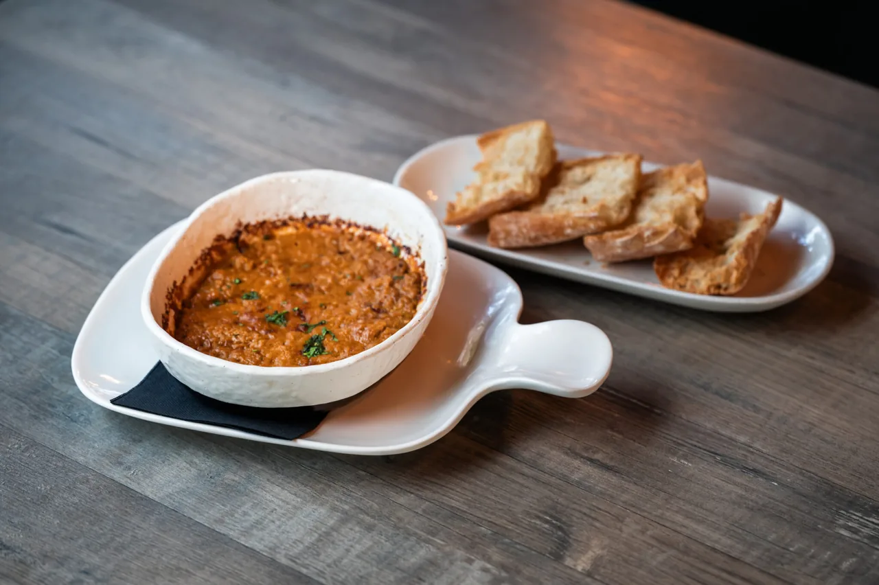A small white handled dish of baked orange-red pate or dip, browned at the rim and scattered with parsley, served on a plate with a black napkin beside a tray of toasted bread slices