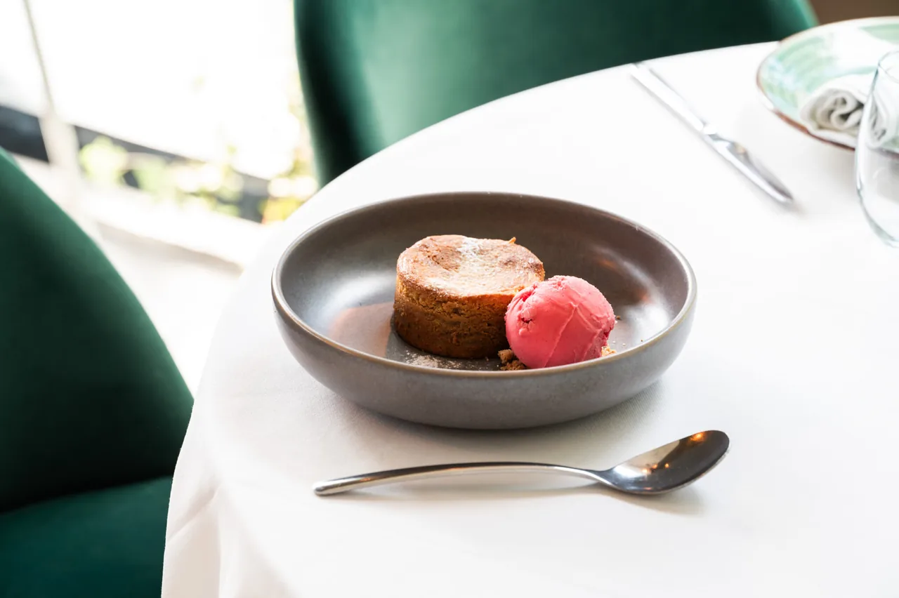 A round baked tart dusted with sugar beside a scoop of bright pink sorbet in a grey bowl, on a white-clothed table with a spoon alongside and a green velvet chair behind