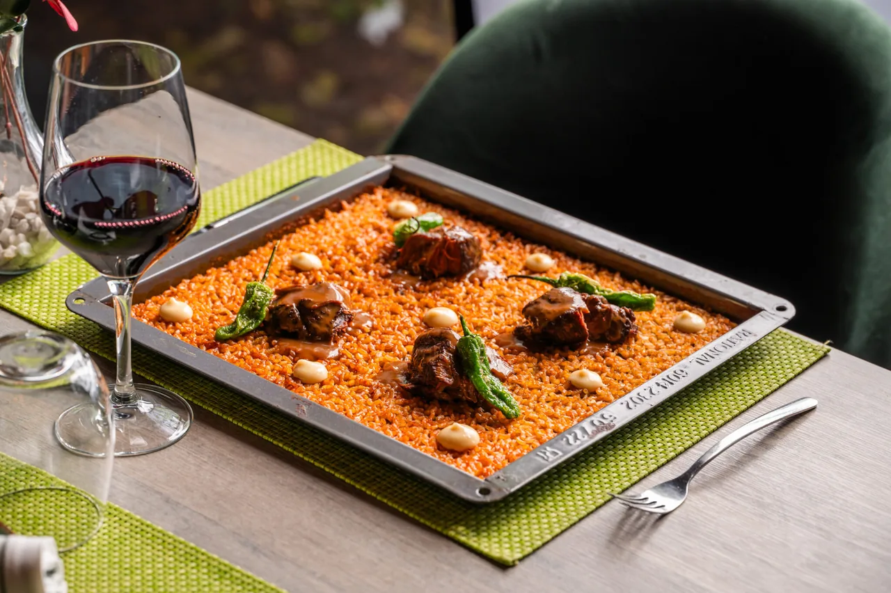 A rectangular metal tray of orange rice topped with chunks of braised meat in dark sauce, blistered padron peppers and dots of pale alioli, on a green placemat with a glass of red wine beside it