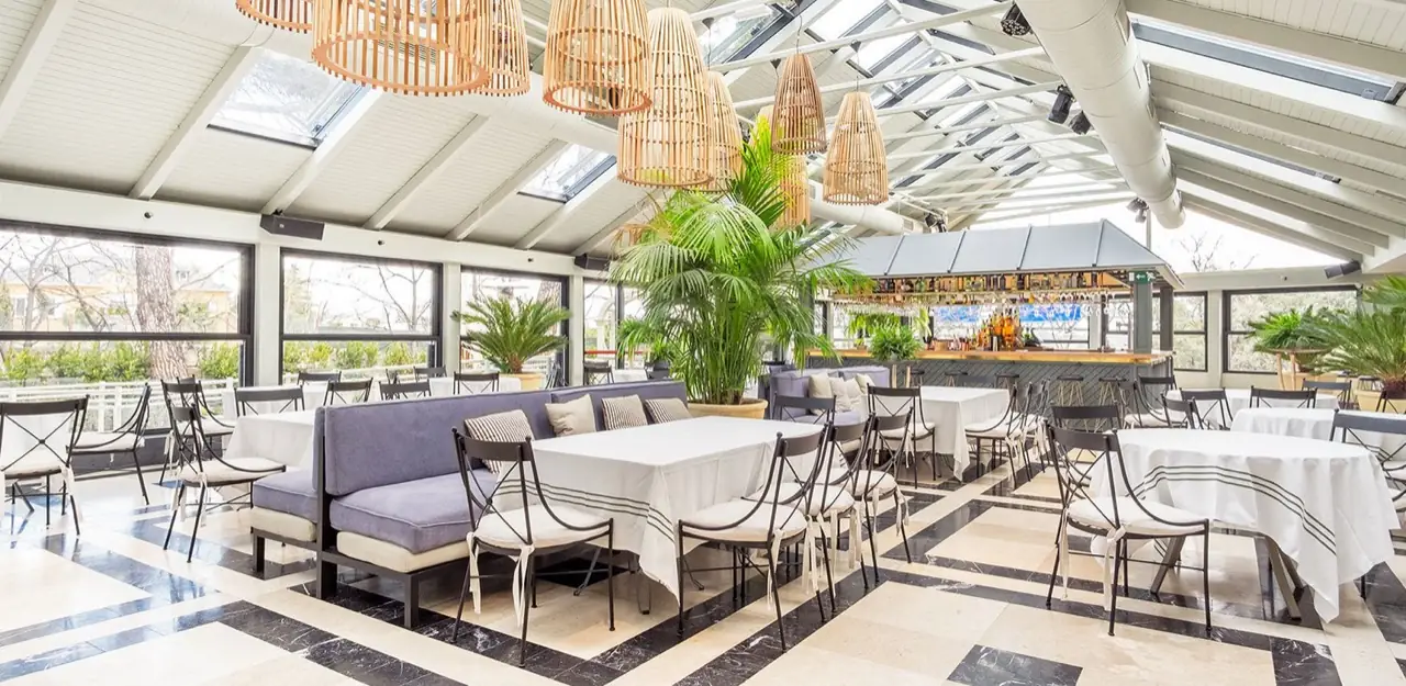 Bright glass-roofed dining room with rattan pendant lamps, white-clothed tables with black metal chairs, a potted palm, and a bar counter in the background.