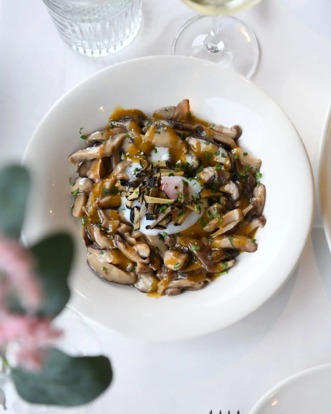 Sauteed mushrooms with a poached egg and shaved truffle in a white bowl on a table set with wine and water glasses