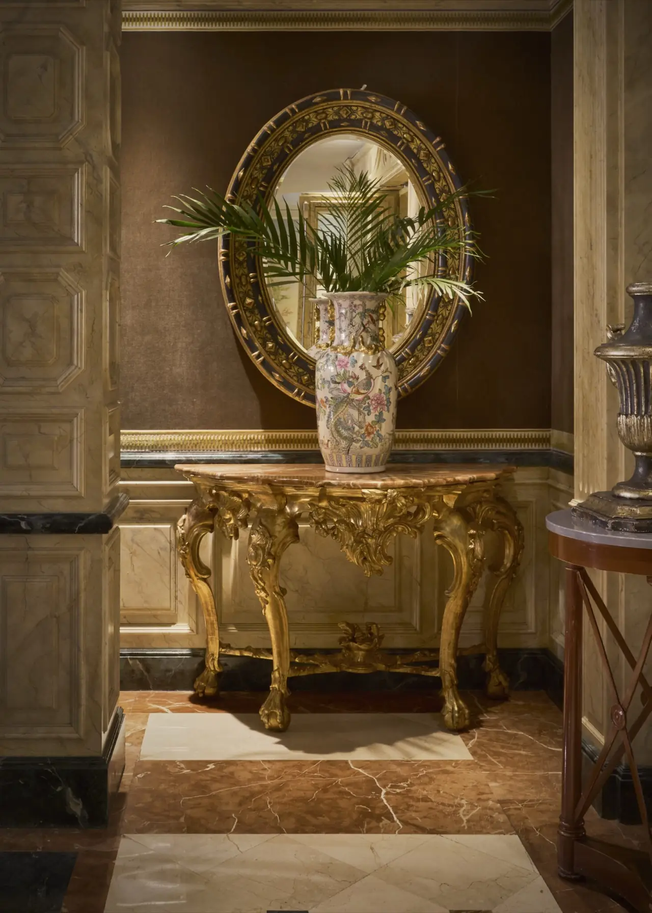 Elegant hotel interior vignette: a gilded rococo console table with a marble top holding a painted floral-and-bird vase, beneath an ornate oval mirror on a brown wall over marble wainscoting