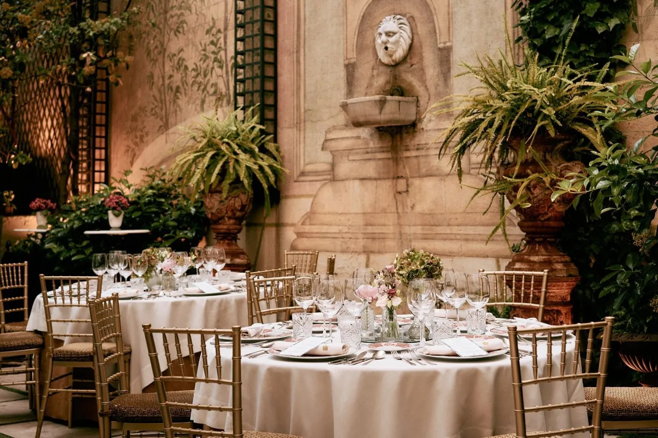 Round tables laid with white tablecloths and flowers in a courtyard garden beside a stone wall fountain