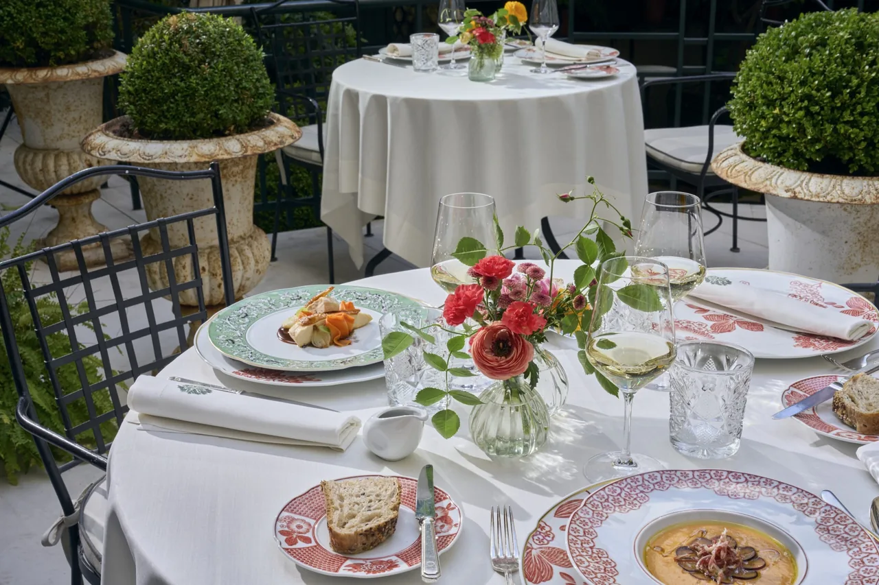 A terrace table with a plated dish, a bowl of soup, wine glasses and a small vase of flowers beside stone planters