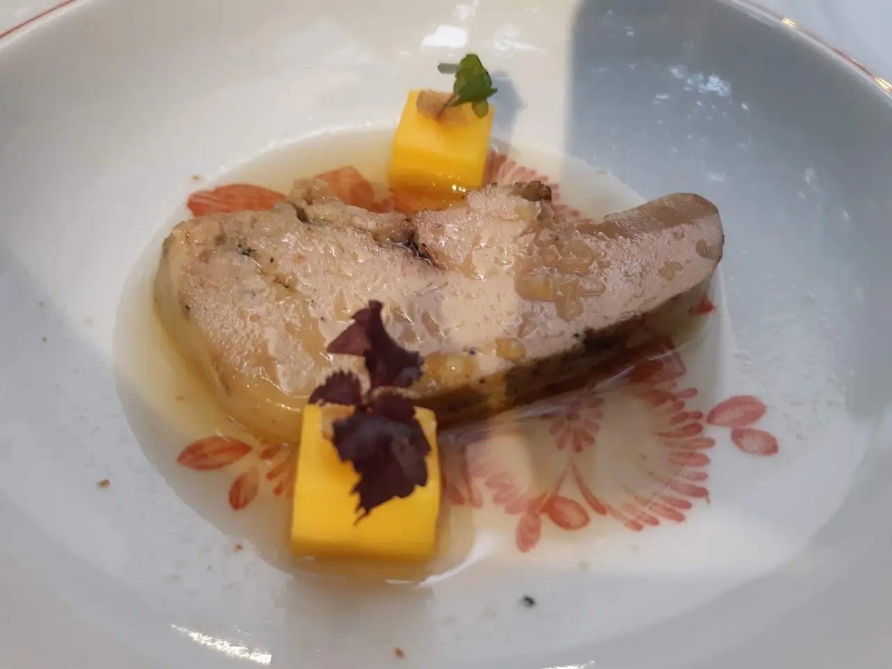 Seared fish fillet with a diced orange garnish and a purple edible flower in a shallow broth.
