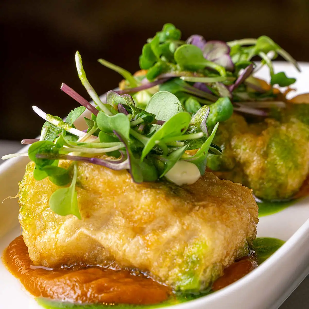 Two battered and fried fish pieces on a white dish over red pepper sauce and green herb oil, piled with red-stemmed microgreens and a dot of mayonnaise