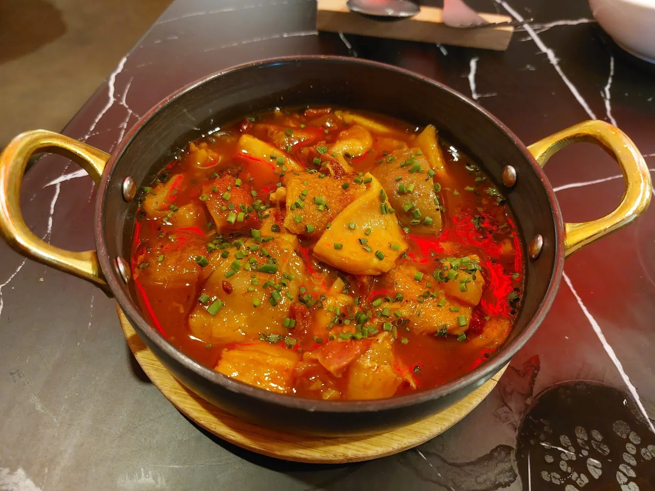 A two-handled black pot of tripe stew in red broth topped with chopped chives