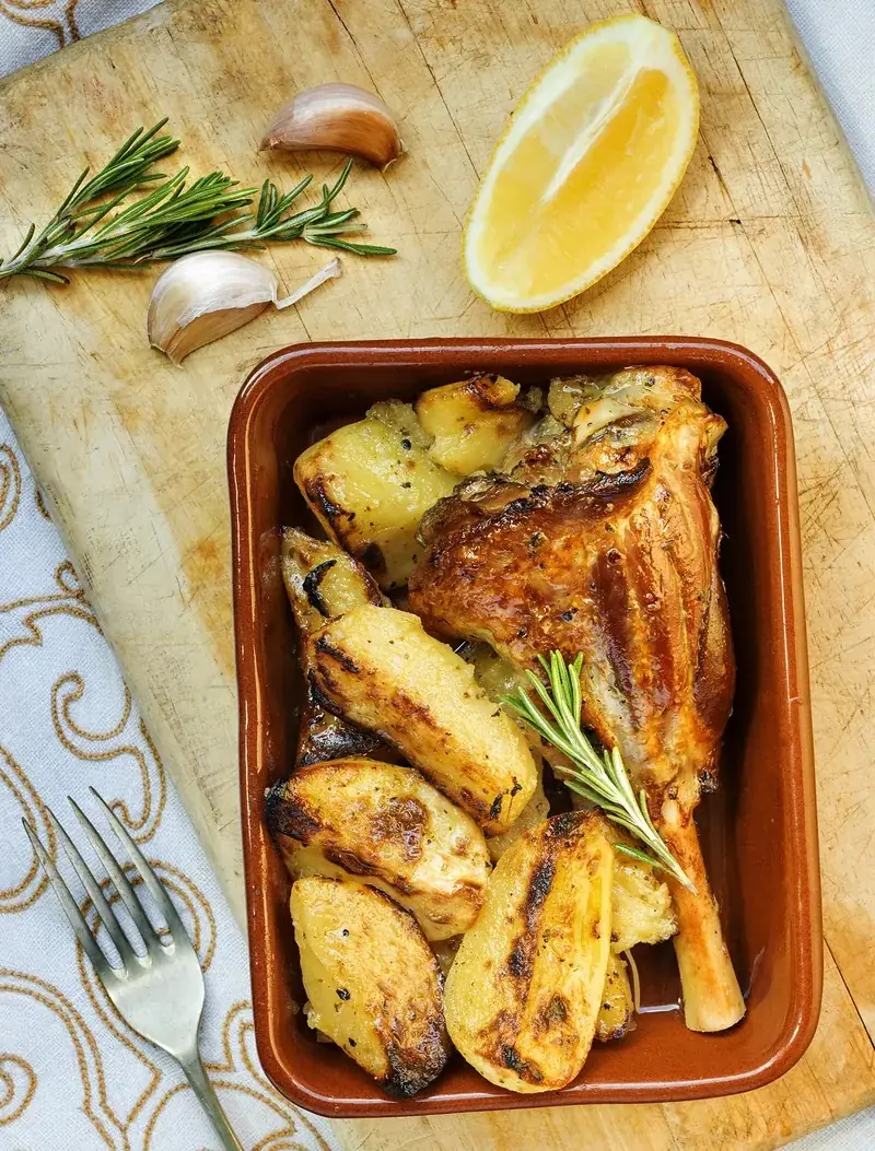Roasted chicken leg with grilled potato wedges in a terracotta dish, with rosemary, garlic, and a lemon wedge, at El Magraner Boig