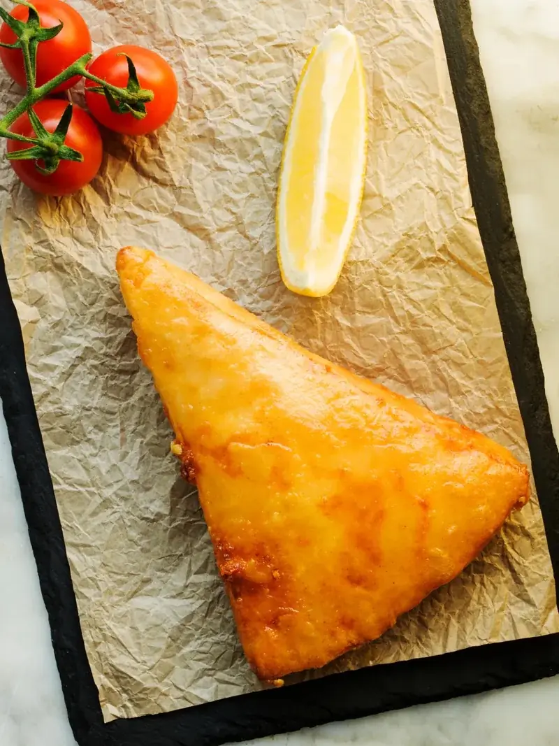 Golden fried pastry triangle served with a lemon wedge and cherry tomatoes on parchment paper, at El Magraner Boig