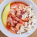 Shrimp ceviche in reddish leche de tigre with avocado slices and cancha corn in a white bowl