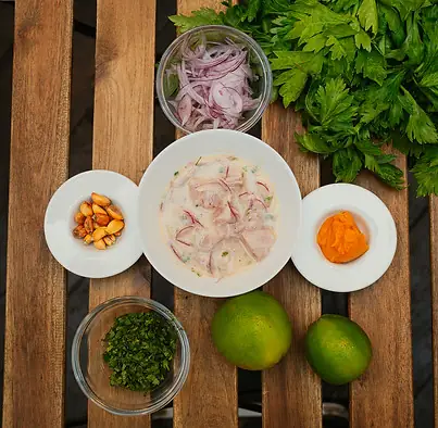 Fish ceviche in a white bowl surrounded by small dishes of red onion, cancha corn, chopped herbs and limes on wooden slats