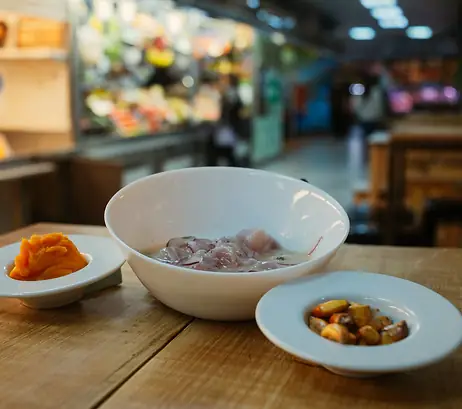 Fish ceviche in a white bowl with sweet potato and side dish on a wooden table inside a market hall