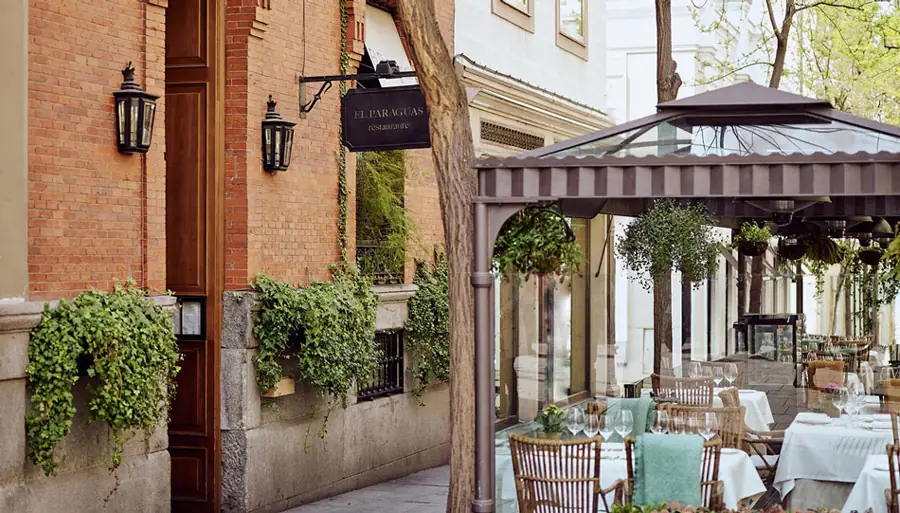 Brick facade with hanging El Paraguas restaurante sign and ivy window boxes beside a covered terrace with set tables