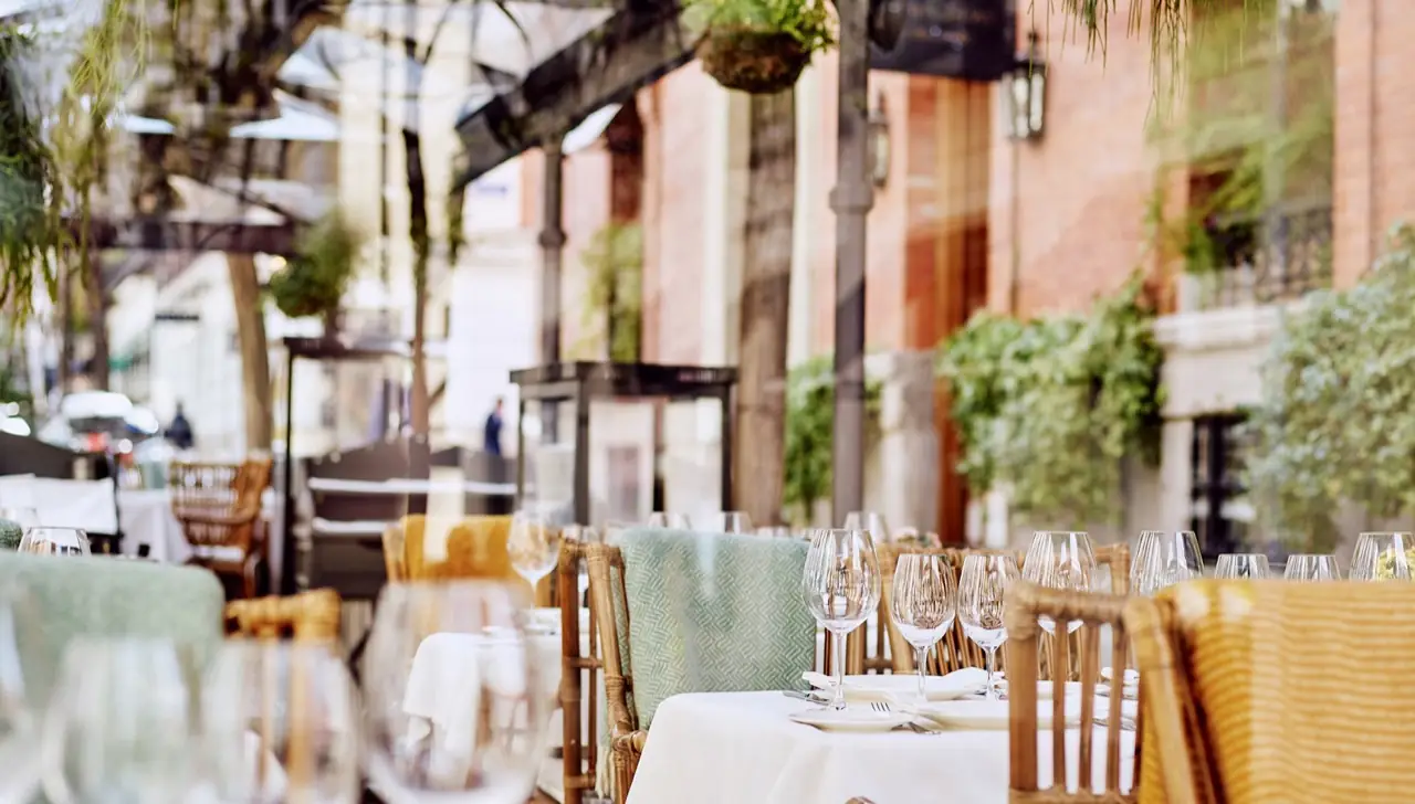 Terrace tables set with wine glasses, white linen and rattan chairs under greenery, brick building behind