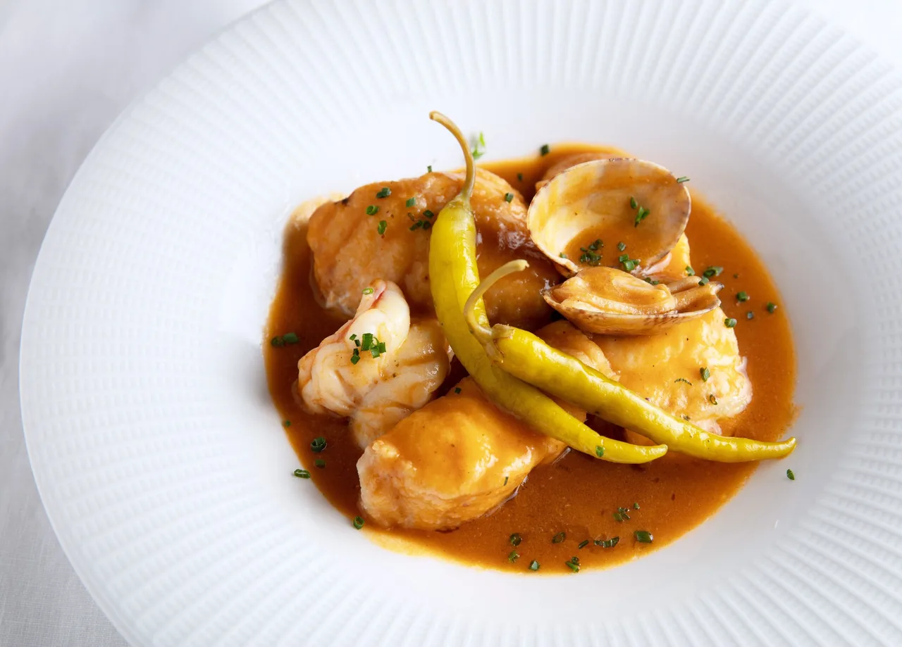 Pieces of seafood with a long green pepper in an orange sauce on a large white plate