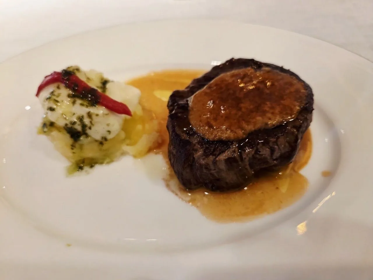 A round dark piece of meat on a white plate beside a small pile of garnish