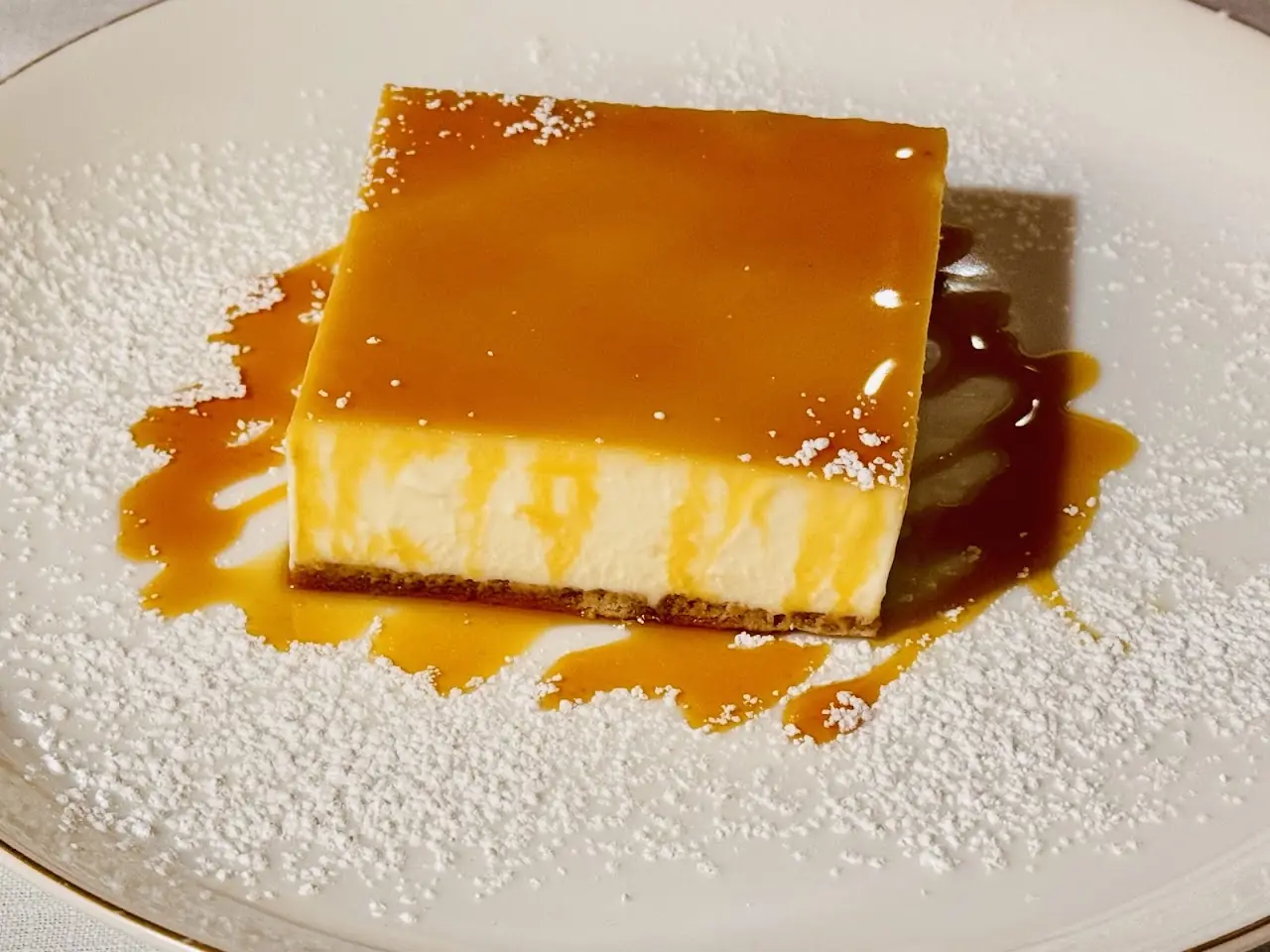 A square caramel flan dessert dusted with powdered sugar on a white plate.