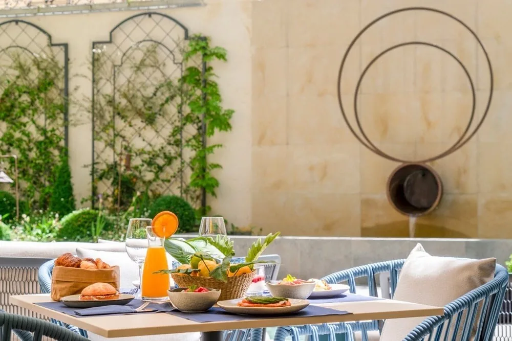 A breakfast spread on a courtyard terrace table with trellis and a stone wall behind