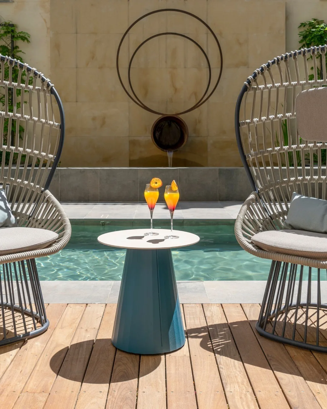 Wicker armchairs and a small table with two orange drinks beside a courtyard pool